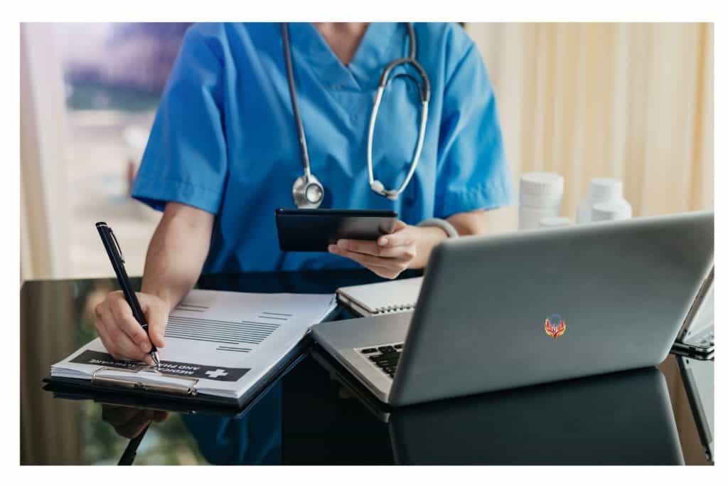 female doctor in blue scrubs taking down information on a page with a laptop sitting on a desk in front of her.