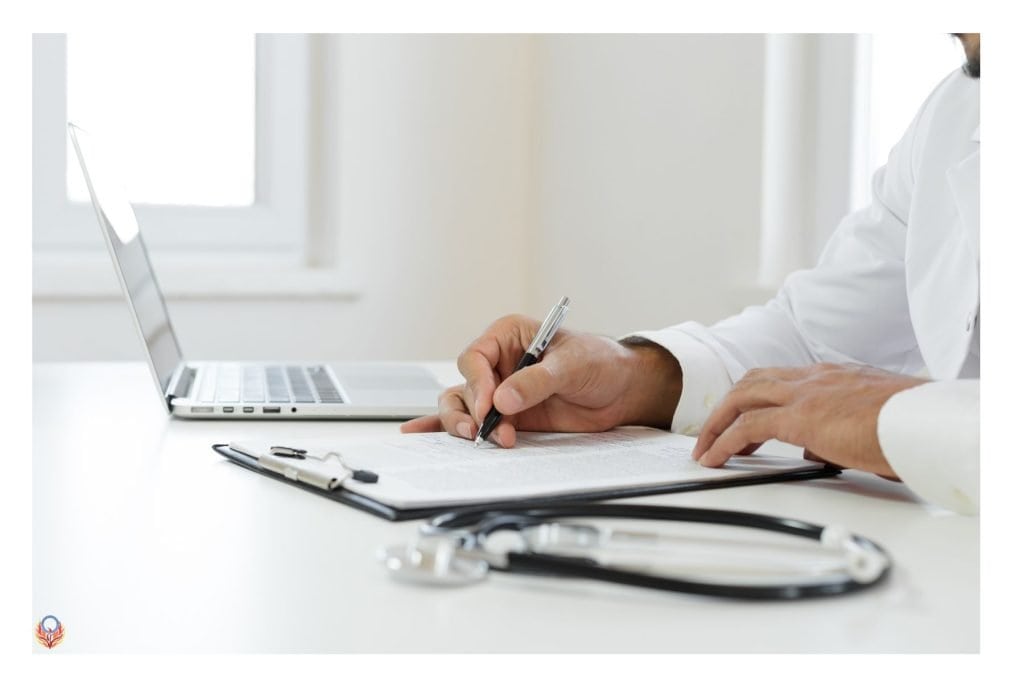 doctor's hands on a note pad writing a prescription. Stethoscope in the foreground and laptop behind his hand.
